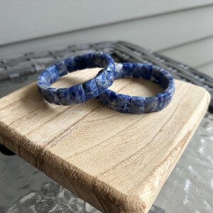 May include: Two blue and white stone bracelets rest on a small, light-colored wooden display stand. The bracelets are made of rectangular-shaped stones, and the stand has rounded edges and black feet. The background is a blurred outdoor setting.