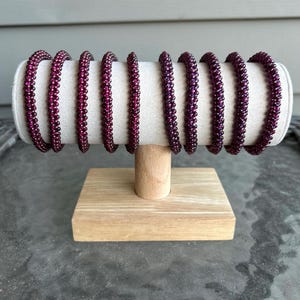 May include: A display of eight beaded bracelets in a rich burgundy color. The bracelets are arranged on a white, cylindrical cushion, supported by a small, natural wood stand. The beads appear to be small and evenly spaced.
