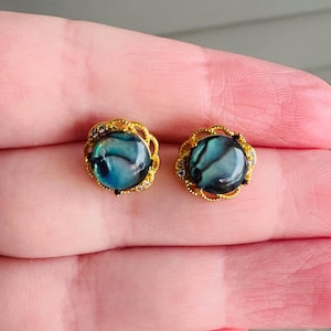 May include: A pair of gold-toned stud earrings. Each earring features a round, iridescent, blue-green stone surrounded by a gold filigree design with small, clear crystals. The earrings are displayed on a light pink background.