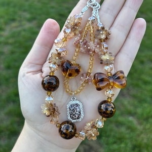 May include: A beaded charm featuring amber and brown leopard print beads, clear crystal flower-shaped beads, and a central rectangular pendant with a leopard print pattern framed in silver-tone metal, all connected by a silver-tone metal clasp.