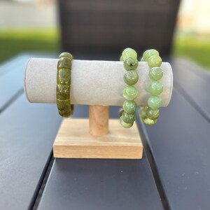 May include: Three green stone bracelets displayed on a light gray, cylindrical jewelry stand with a wooden base. The bracelets feature varying bead shapes and shades of green, from olive to light green. The stand is on a dark gray surface.