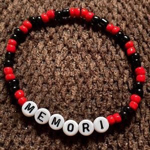 May include: A beaded bracelet with alternating red and black beads. White round beads spell out the word "MEMORI". The bracelet is on a brown, textured surface.