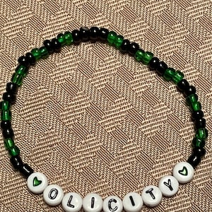 May include: A beaded bracelet with black and green beads. The bracelet spells out "OLICITY" in white letter beads, with green heart beads at each end. The bracelet is on a brown patterned surface.