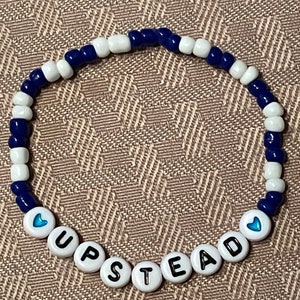 Puede incluir: Una pulsera de cuentas con cuentas blancas y azul marino. La pulsera deletrea "UPSTEAD" con cuentas de letras blancas. Dos de las cuentas de letras tienen un símbolo de corazón azul. La pulsera está sobre una superficie marrón texturizada.