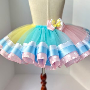 May include: A rainbow tulle tutu skirt with layers of pink and blue ribbon, some with white polka dots. The skirt has a yellow, orange, green, blue, and pink ombre effect.