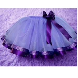 May include: A lavender tutu skirt with a purple satin ribbon bow and trim. The skirt has layers of tulle and a glittery band at the hem. The bow and trim are a darker shade of purple.