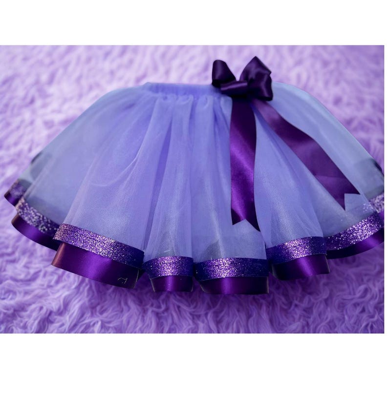 May include: A lavender tutu skirt with a purple satin ribbon bow and trim. The skirt has layers of tulle and a glittery band at the hem. The bow and trim are a darker shade of purple.