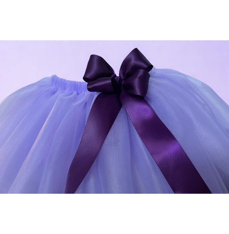 May include: A light purple tutu skirt with a dark purple satin bow and matching ribbon. The skirt is made of layers of soft tulle, creating a full and fluffy appearance. The bow is centered at the waist.