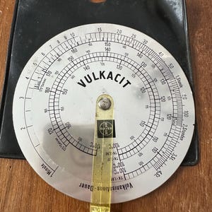 May include: A vintage Vulkacit circular slide rule with a yellow plastic arm and black case. The slide rule features temperature and time scales, with the Bayer logo visible. The device is designed for calculating vulcanization processes.