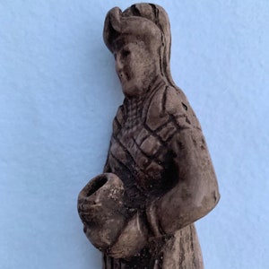May include: A brown ceramic figurine of a person wearing a hat and holding a round object in their hand.