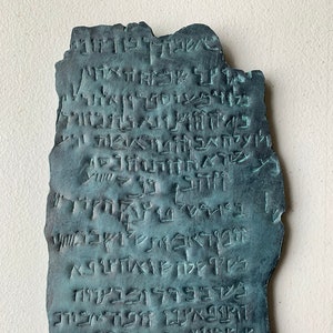 May include: A close-up of a dark green, rectangular, stone tablet with ancient Hebrew writing carved into the surface. The tablet has a rough, textured surface and is slightly chipped on the edges.