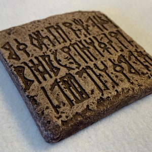 May include: A square, brown, stone-like coaster with a pattern of carved runes. The runes are in a dark brown color and are carved into the surface of the coaster.