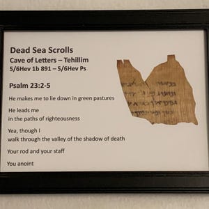 May include: A framed print of a Dead Sea Scroll fragment with the text of Psalm 23:2-5. The text reads: "He makes me to lie down in green pastures. He leads me in the paths of righteousness. Yea, though I walk through the valley of the shadow of death. Your rod and your staff. You anoint."
