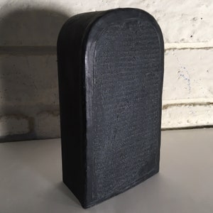 May include: A black, rectangular, stone-like object with a raised, arched top. The surface is covered in a dense pattern of small, raised characters.