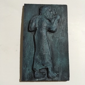 May include: A dark blue wooden plaque with a raised relief sculpture of a person in a long robe. The sculpture is painted in shades of blue and green.