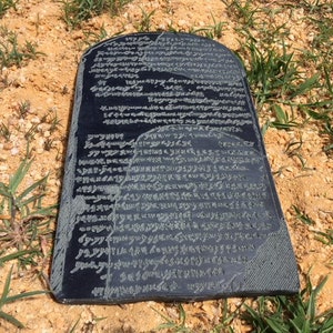 May include: A black stone tablet with ancient writing carved into its surface. The tablet is lying on a bed of sand and grass.