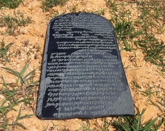 May include: A black stone tablet with ancient writing carved into its surface. The tablet is lying on a bed of sand and grass.