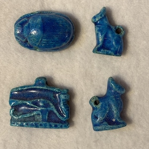 May include: Four blue glazed ceramic charms including a scarab beetle, two small animal figures, and a rectangular piece featuring an Eye of Horus symbol. Each piece has a smooth, glossy finish and a small hole for stringing or attachment.