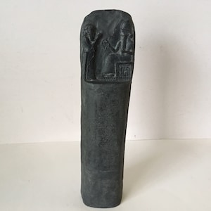 May include: A black stone stele with cuneiform script and a relief carving of two figures. The figures are a seated goddess holding a staff and a standing figure holding a cup.