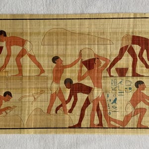May include: A vintage papyrus scroll depicting a scene of ancient Egyptian workers carrying large blocks of stone. The scroll is beige with brown and red figures. The scene is framed by a border of green leaves and a blue square with a stylized flower.