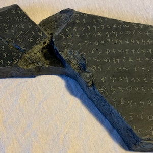May include: A dark gray stone fragment with etched text. The stone is broken into two pieces, with the text appearing to be an ancient script. The surface of the stone is rough and textured, with the carved letters in a lighter color.