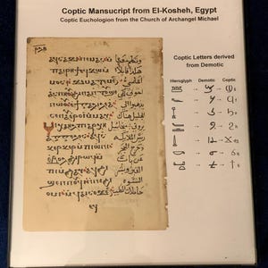 May include: A framed display featuring a Coptic manuscript from El-Kosheh, Egypt, with text in ancient script. The display includes a section comparing Coptic letters derived from Demotic, with hieroglyphs and their Coptic equivalents. The text is in black ink on aged paper.