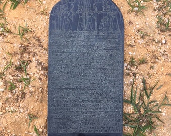 May include: A black stone slab with ancient Egyptian hieroglyphics carved into the surface. The slab is shaped like a rounded rectangle and has a raised border around the edges. The hieroglyphics depict a scene of people offering gifts to a deity.