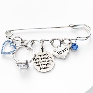 May include: Silver bridal brooch with charms including a heart with the word "Bride", a ring, a heart with rhinestones, and a round charm with the text "my little girl yesterday my friend today my daughter forever".