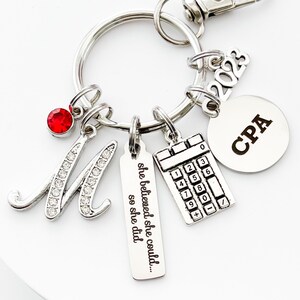 May include: Silver keyring with a red rhinestone, a letter M with rhinestones, a charm with the text "she believed she could... so she did", a calculator charm, a charm with the text "CPA", and a charm with the year 2023.