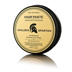 Puede incluir: Un tarro negro con tapa dorada que contiene pasta para el cabello. El tarro tiene una etiqueta dorada con el texto "The Golden Spartan" y "Handmade Petroleum Free" en negro. La etiqueta también enumera los ingredientes y el tamaño del tarro, que es de 125g/4.4oz.
