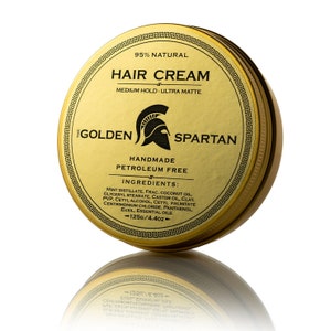 Puede incluir: Contenedor dorado con una imagen en blanco y negro de un casco espartano y el texto "The Golden Spartan" y "Handmade Petroleum Free". El contenedor está etiquetado como "Hair Cream" y enumera los ingredientes, "95% Natural", "Medium Hold - Ultra Matte", y "125g/4.4oz".