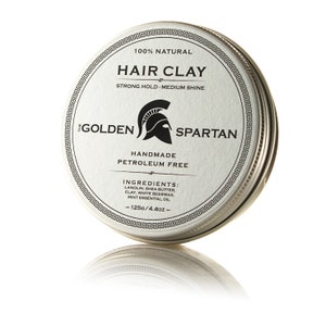 Puede incluir: Una lata plateada con una etiqueta blanca que dice "100% NATURAL HAIR CLAY STRONG HOLD-MEDIUM SHINE THE GOLDEN SPARTAN HANDMADE PETROLEUM FREE INGREDIENTS: LANOLIN, SHEA BUTTER, CLAY, WHITE BEESWAX, MINT ESSENTIAL OIL - 125G/4.4OZ -" con un gráfico de casco espartano.