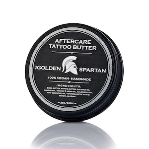 May include: A round, black tin of "AFTERCARE TATTOO BUTTER" with a white logo of a Spartan helmet. The label also states "100% VEGAN - HANDMADE" and lists ingredients like shea butter and coconut oil. The tin contains 25g / 0.9oz of product.