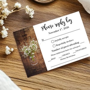 May include: A rectangular wedding RSVP card with a white background and a dark wood grain panel on the left. The card features a graphic of a mason jar with baby's breath flowers and text that reads Please reply by November 1st, 2026.