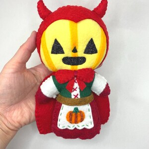 May include: A handcrafted felt plush doll featuring a yellow pumpkin-shaped head with black glittery triangular eyes and a mouth. The doll wears a red hooded cape with horns, a green bodice with a red bow, and a white apron embroidered with a pumpkin.