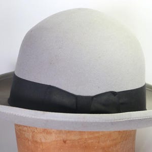 May include: A light gray fedora hat with a black band. The hat has a rounded crown and a wide brim. The hat is displayed on a wooden hat block.