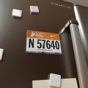 May include: A white refrigerator magnet with a colorful Chicago Marathon race bib. The bib is orange and blue with the text "Bank of America Chicago Marathon" and "N 57640" with the date "October 9, 2022".
