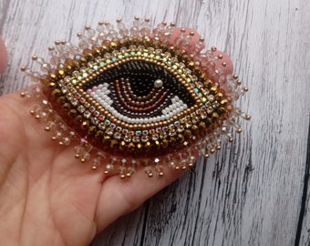May include: A beaded eye brooch with a black pupil, white iris, and brown and gold accents. The eye is surrounded by a fringe of clear beads.
