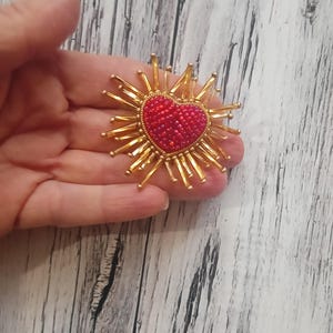 May include: A heart-shaped brooch with a red beaded center and gold-colored rays radiating outwards. The brooch is held in a hand against a light wood-grain background. The brooch is a decorative accessory.