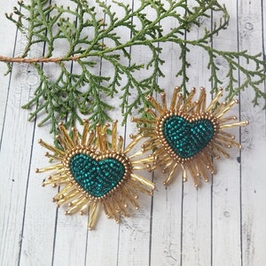 May include: A pair of gold beaded earrings with a green heart-shaped center. The earrings are shaped like a sunburst with gold beads radiating out from the center.