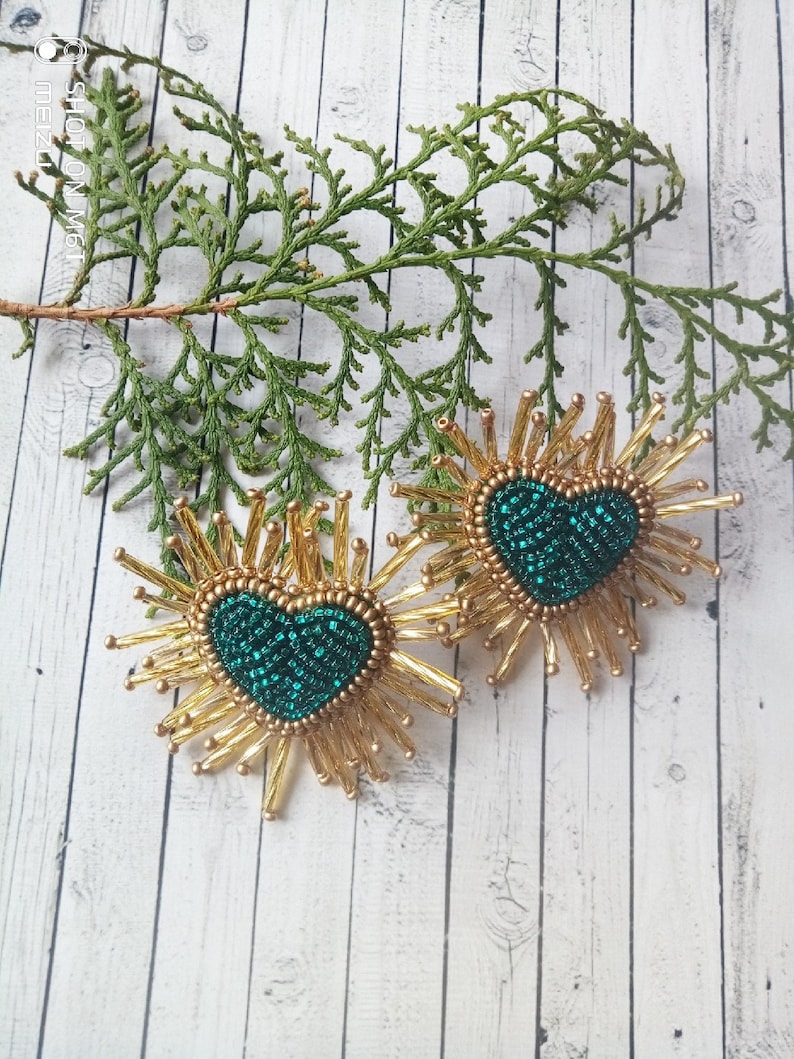 May include: A pair of gold beaded earrings with a green heart-shaped center. The earrings are shaped like a sunburst with gold beads radiating out from the center.