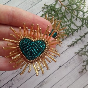 May include: A pair of gold beaded earrings with a green heart-shaped center. The earrings are made of small gold beads that are arranged in a sunburst pattern around the heart.