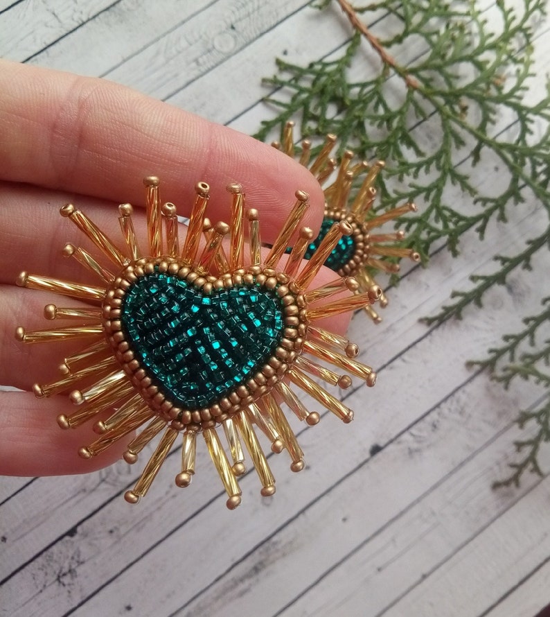 May include: A pair of gold beaded earrings with a green heart-shaped center. The earrings are made of small gold beads that are arranged in a sunburst pattern around the heart.