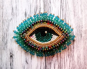 May include: A handcrafted eye-shaped brooch with intricate beadwork. The design features a black pupil, a green iris, and a gold-colored outline, surrounded by teal and iridescent beads. The brooch is set against a light wood grain background.