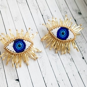 May include: Two gold beaded evil eye charms with blue and white centres. The charms are on a white wooden background.