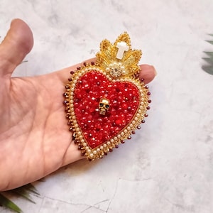 Op de afbeelding: Een rood hart van kralen met een gouden schedel en een gouden kruis bovenop. Het hart is omgeven door een gouden kralenrand en witte parels.