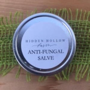 May include: A round, silver tin with a white label that reads "HIDDEN HOLLOW FARM" and "ANTI-FUNGAL SALVE" in black text. The tin sits on a piece of green burlap, against a wooden background.