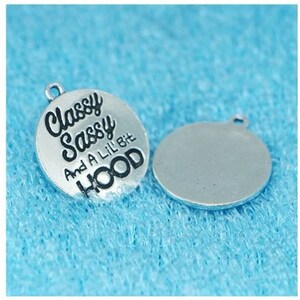 May include: Two silver-toned metal charms. One charm is round and features the text "Classy Sassy And A Lil Bit Hood" in black lettering. The other charm is blank.