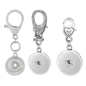 May include: Three silver-toned snap-on charm keychains with different styles of clasps. Each keychain has a round, silver-toned charm with a small hole in the center.