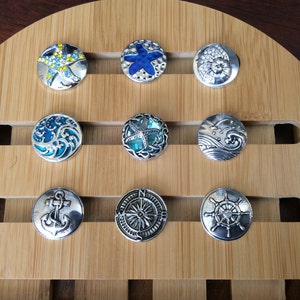 May include: A wooden display board with 10 round, silver-toned snap jewelry charms. The charms feature various nautical themes, including seashells, starfish, anchors, and a compass.